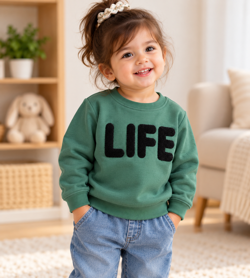 Smiling toddler girl wearing a green fleece winter sweatshirt with black textured LIFE lettering, round neck and ribbed cuffs, paired with blue jeans – front view.