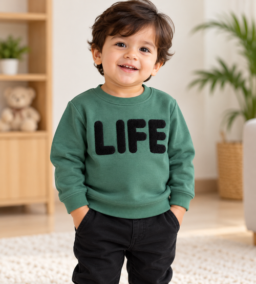Smiling toddler boy wearing a green fleece winter sweatshirt with black textured LIFE lettering, round neck and ribbed cuffs – front view.