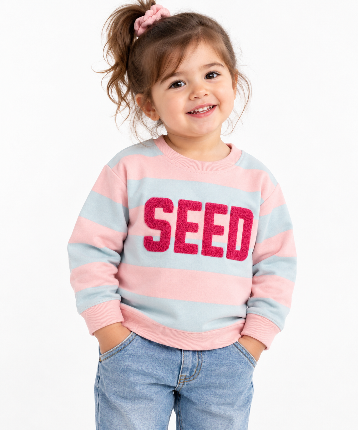Smiling toddler girl wearing a pink and light blue striped fleece sweatshirt with textured SEED lettering, round neck and ribbed cuffs, paired with blue jeans – front view.