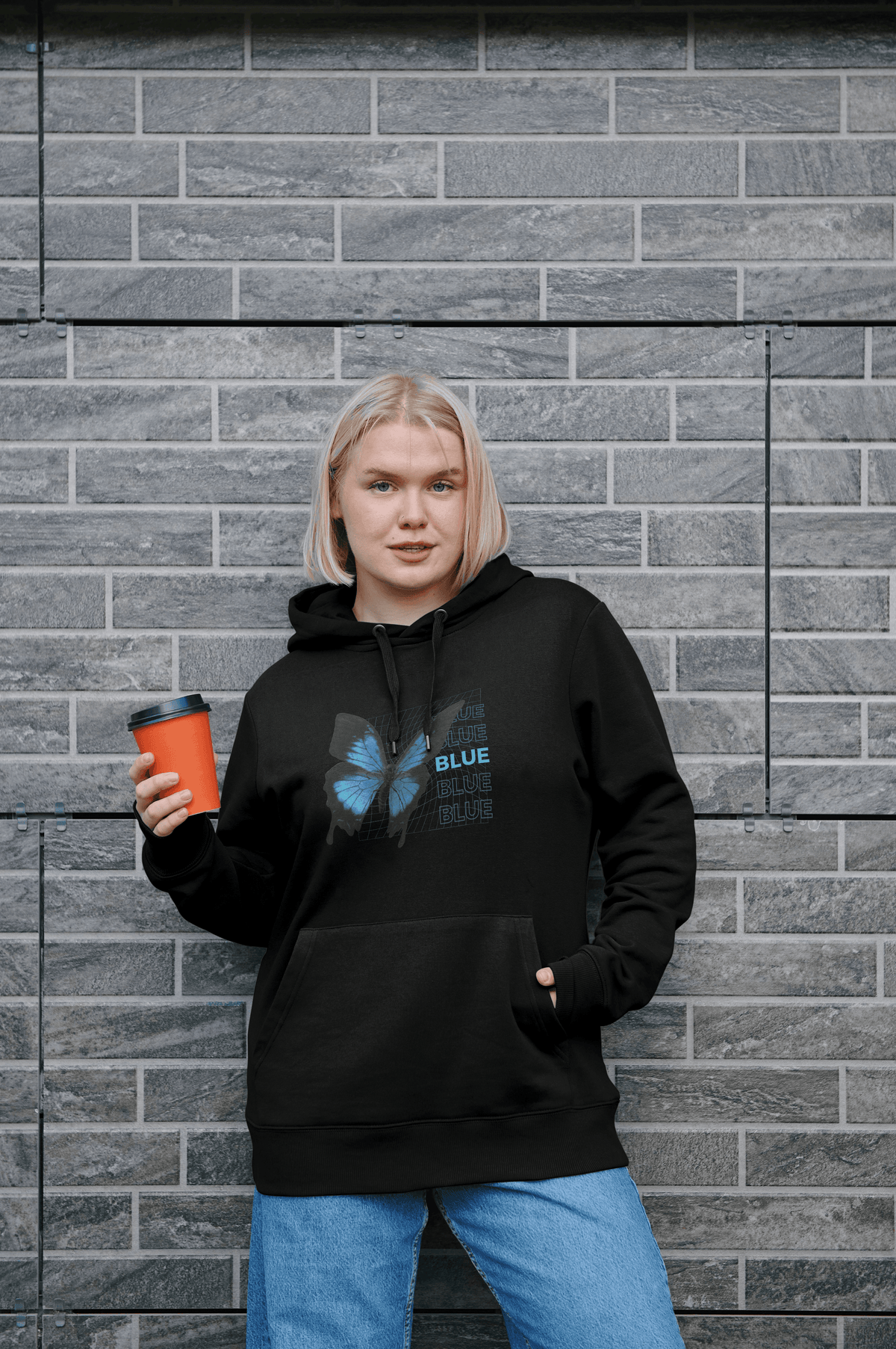 A woman with blonde hair stands against a gray brick wall, wearing a black hoodie with a blue butterfly graphic and the text 'BLUE BEYOND DARKNESS' on the front, and holding an orange coffee cup.