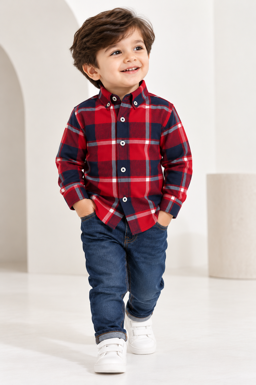Smiling toddler boy wearing a red and navy checked flannel shirt with blue jeans and white shoes in a bright indoor studio.