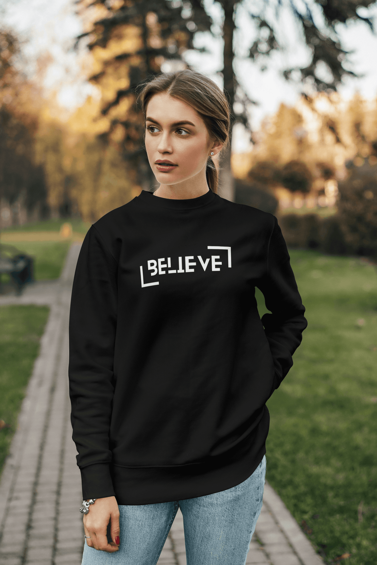 A woman wearing a black crewneck sweatshirt with the word 'BELIEVE' printed in white geometric letters, standing on a paved path in a park with trees in the background.