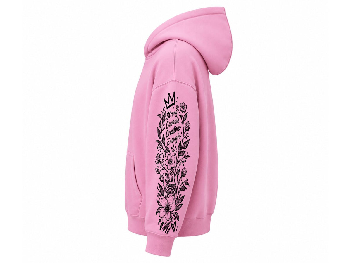 A pink pullover hoodie shown from the side, featuring a black graphic print on the sleeve with a crown, floral elements, and the words 'She Is Comfortably Alone' on a plain white background.